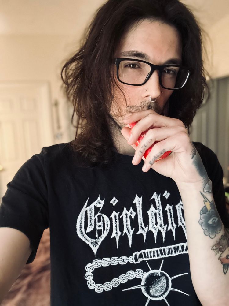 selfie of my in my room wearing a black shirt that says Girldick with a morningstar under it. I'm holding a vape in my hand in front of my mouth. my hair is parted in the middle and resting at my shoulders. part of my hand tattoo is visible, and you can see my storm cloud tattoo and part of my fox farmer