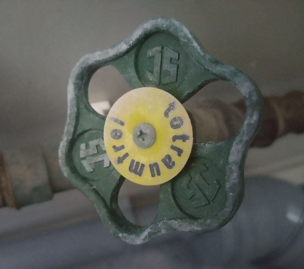 Grünes Handrad am Absperrventil einer Wasserleitung mit gelbem Schild in der Mitte.  Darauf kreisförmig geschrieben das Wort „totraumfrei“.