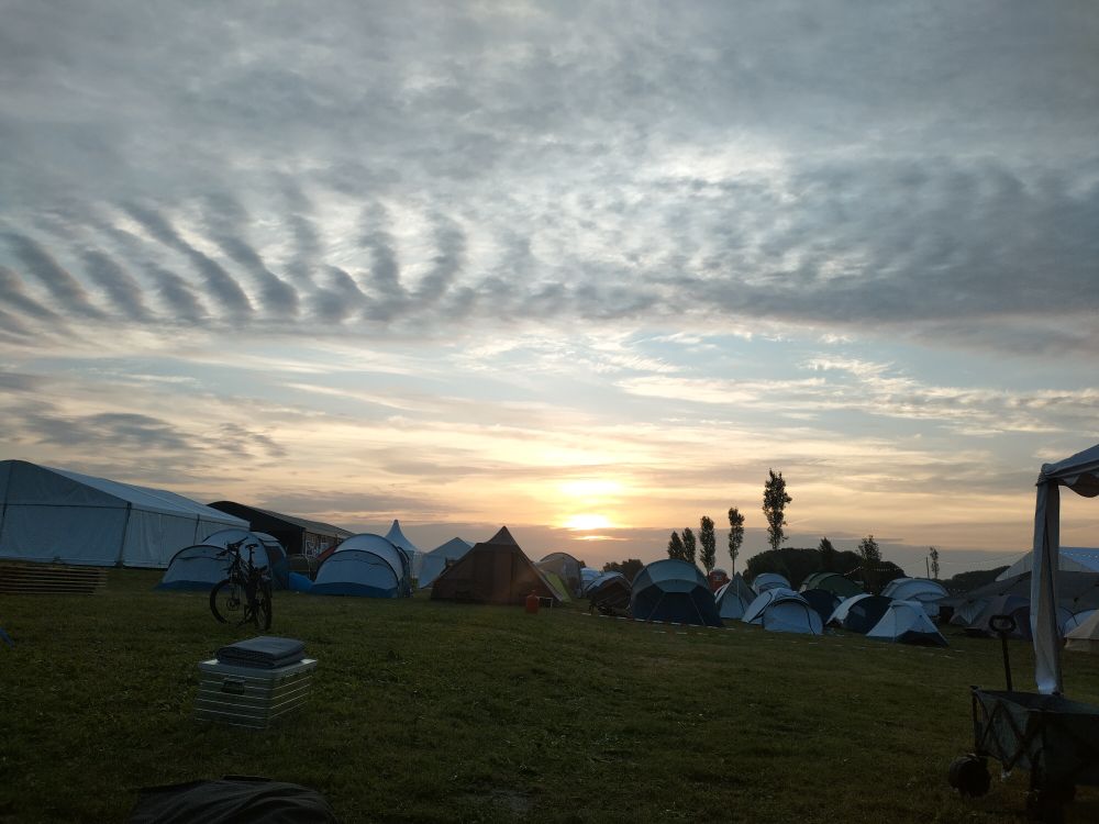 Zelte im Halbdunkel, dahinter müht sich die Sonne redlich durch geschichtete Wolken.  Weiter oben ein vertikales Tigerstreifen-Wolkenmuster.