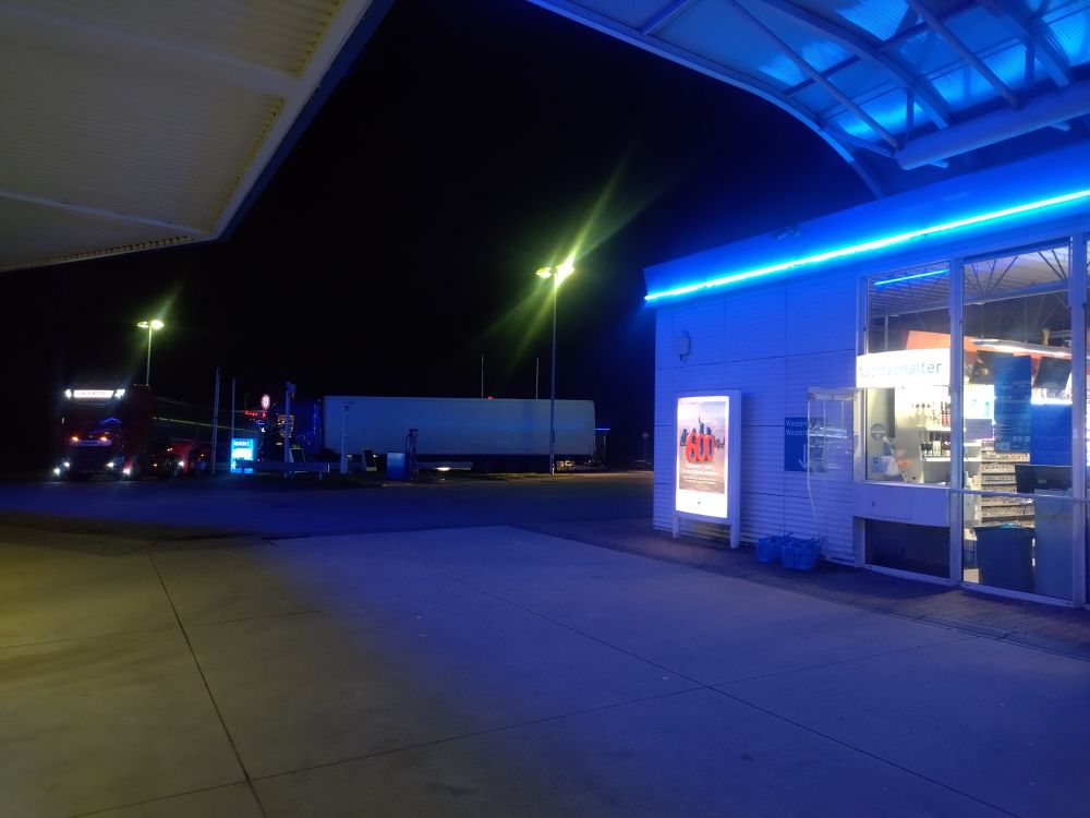 Rechts ein Tankstellengebäude mit blauem Leuchtstreifen, im Hintergrund einige parkende LKW.  Der Nachtschalter ist überbelichtet, aber zu erkennen.