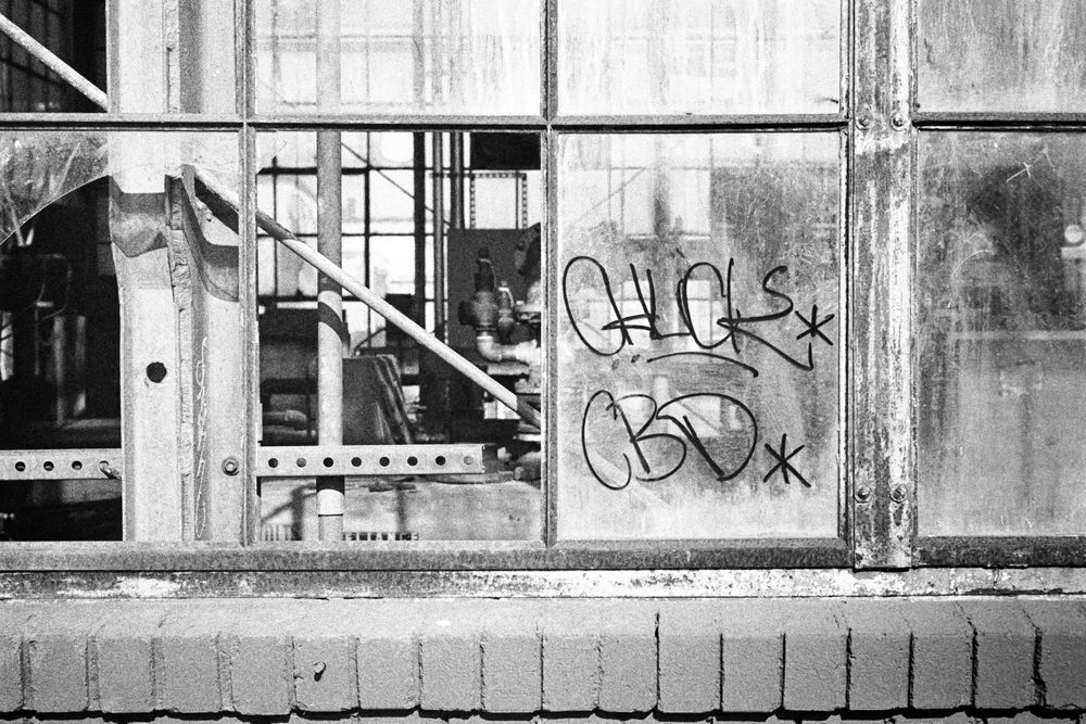 A black and white photograph of a partially broken and partially graffitied window in front of an abandoned industrial space