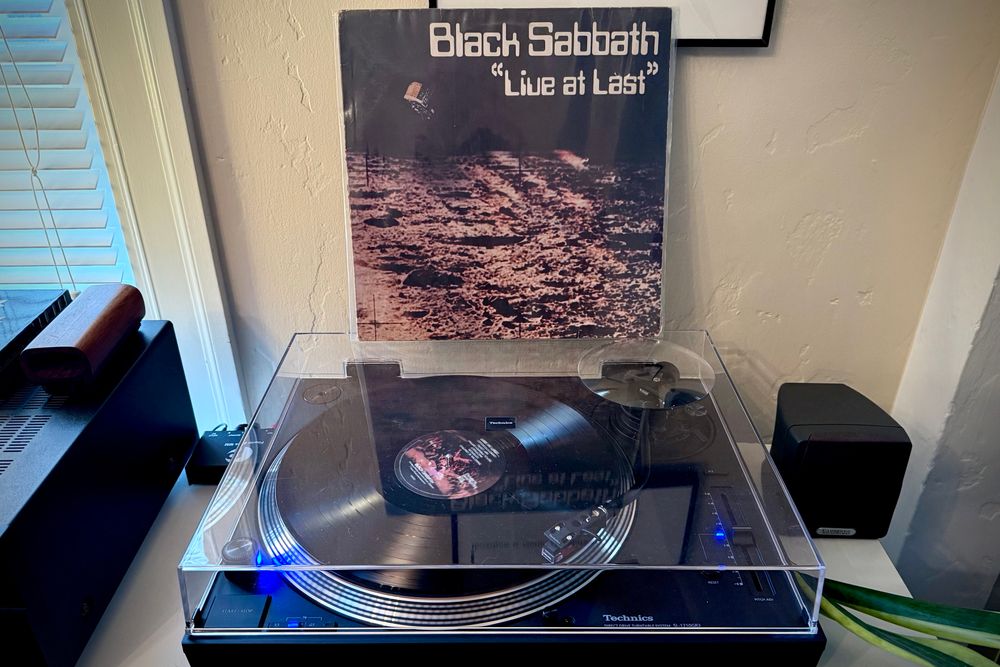 A black vinyl record spins on a black turntable. The record sleeve stands up on the turntable’s closed dust cover behind the platter. 