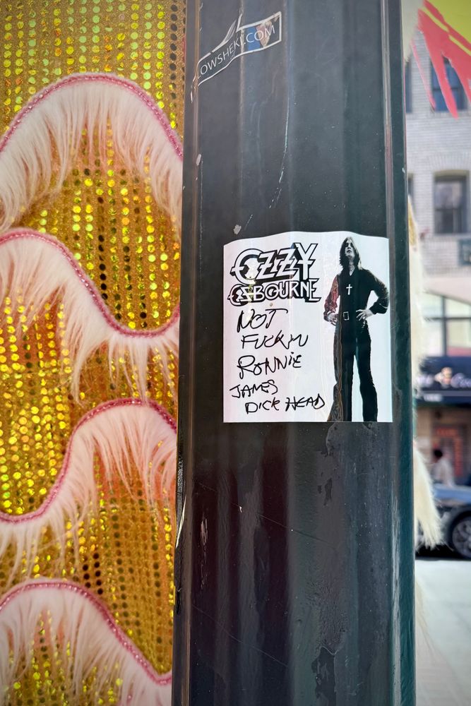 A sticker on a light pole, gold sequined and pink ruffled curtain behind it. The sticker features a photo of young Ozzy Osbourne and says, “Ozzy Osbourne Not Fuckin Ronnie James Dickhead”