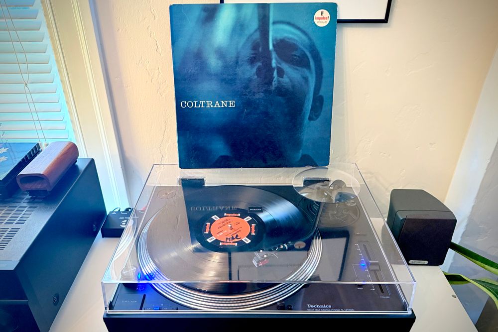 A black vinyl record spins on a black turntable. The record sleeve stands up on the turntable’s closed dust cover behind the platter. 