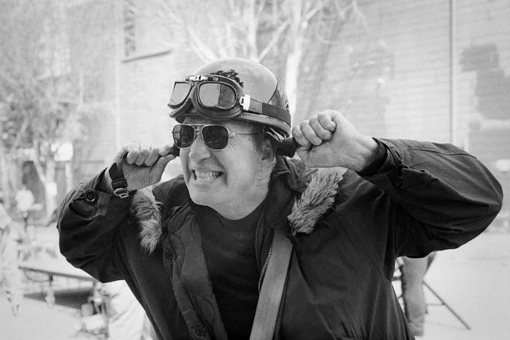 A black & white photograph of a person in a motorcycle helmet and sunglasses, gripping and pulling on the helmet straps while grimacing 