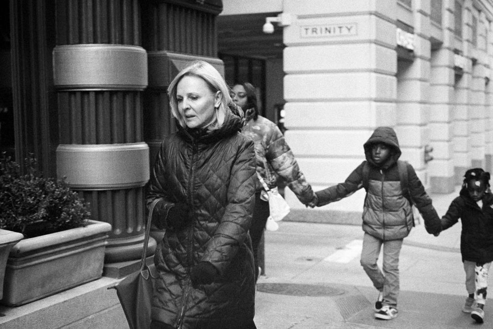 A black and white photo of a person a puff coat, followed by a family of three holding hands, also wearing winter coats
