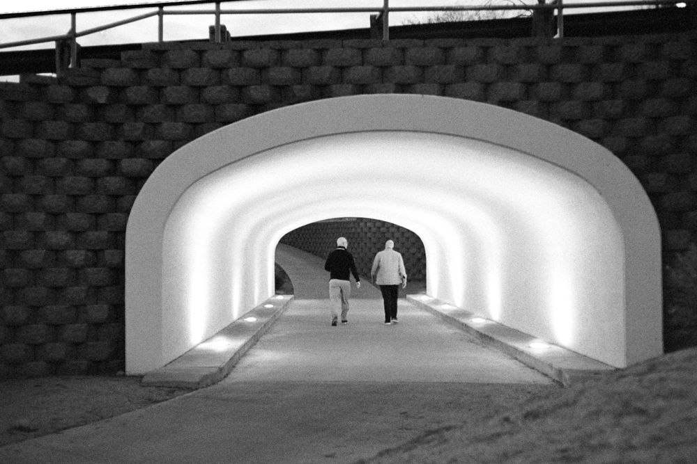 A black & white photograph of two people walking away through a well lit tunnel, surrounded by a darkened structure 