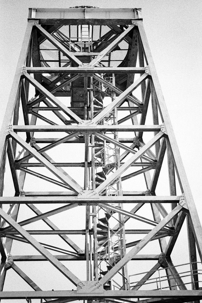 A black & white photograph looking up at an abandoned steel-frame tower with a spiral staircase 