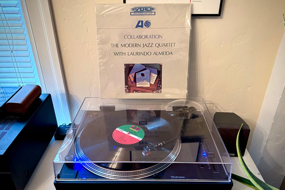A black vinyl record spins on a black turntable. The record sleeve stands up on the turntable’s closed dust cover behind the platter. 