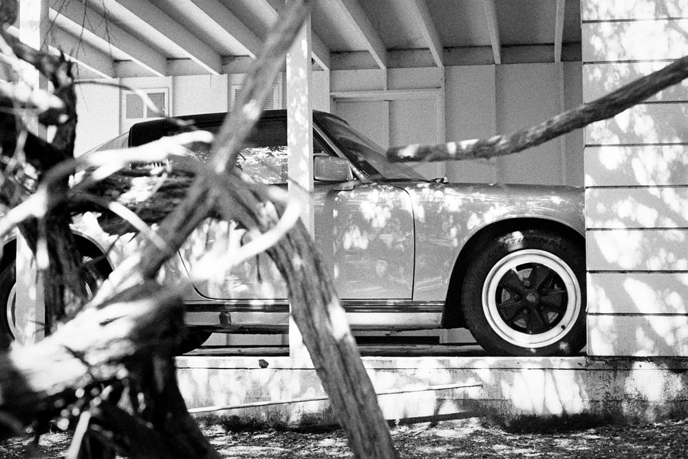 A black and white photo of a classic sports car in a covered driveway, partially obscured by foreground trees 