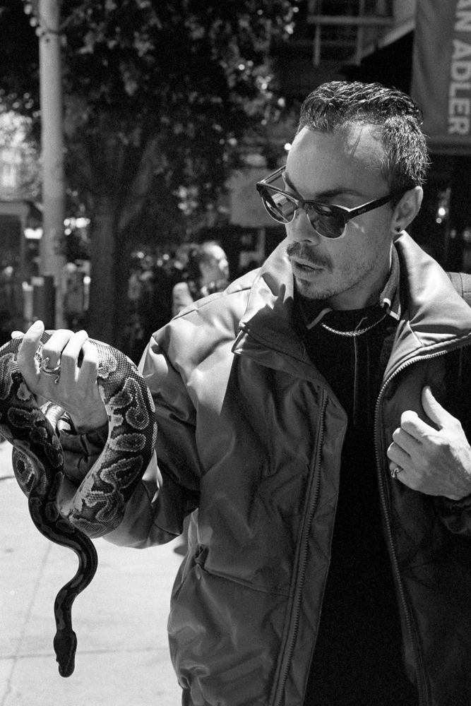 A black and white photograph of a person in sunglasses and a light puff jacket looking at the snake that he’s holding in his right hand. 