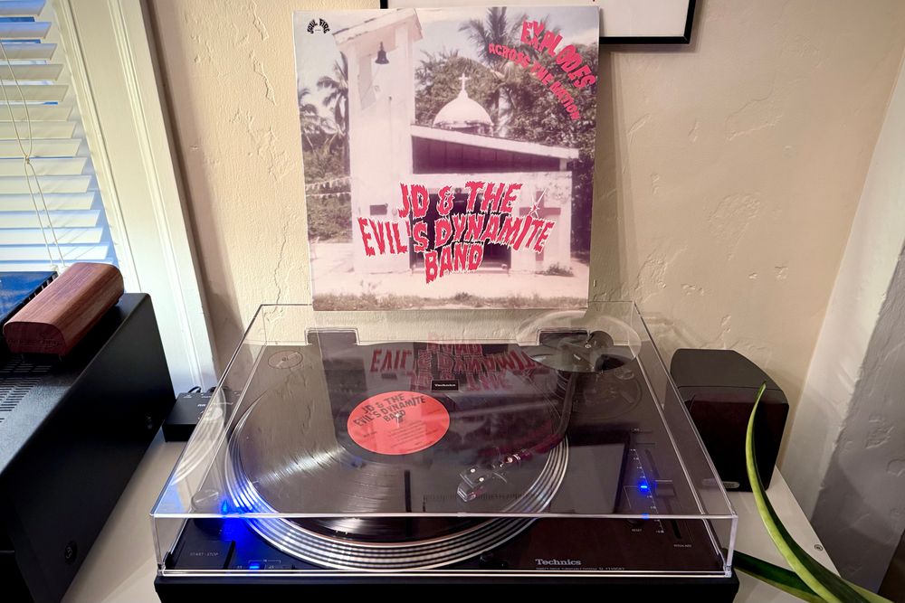 A black vinyl record spins on a black turntable. The record sleeve stands up on the turntable’s closed dust cover behind the platter. 