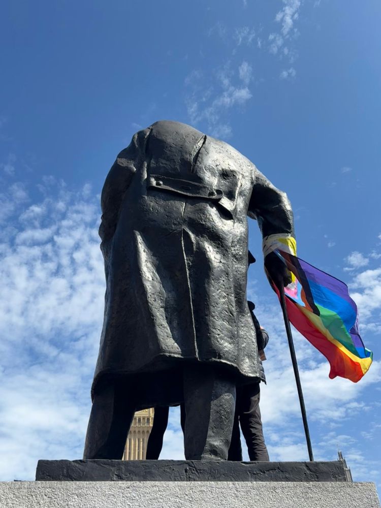 The statue of Churchill in Parliament Square, seen from behind, with protestors’ legs visible on the other side of the statue and a progress pride flag fluttering from his stick against a blue sky with a dusting of cloud.