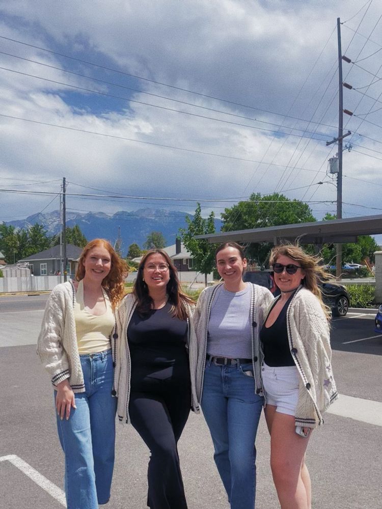 four women standing all wearing off white cardigans in a parking lot with houses, power lines, mountains, and clouds in the background. from left to right the first woman has red hair and is wearing blue jeans, a yellow top. the second woman is a brunette with glasses and is wearing a black top and black pants. the third woman is a brunette with her hair pulled back and is wearing a blue top and blue jeans with a black belt. the fourth woman is a dirty blonde with sunglasses and is wearing a black top and white shorts. 