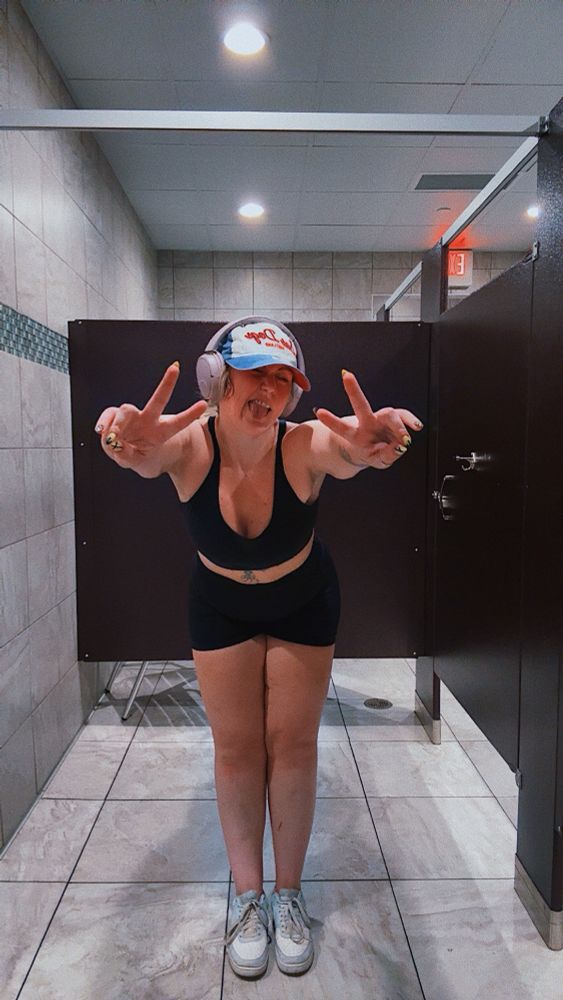 a woman standing in a locker room stall while bent at the waist making peace signs and a silly face with her tongue out. she is wearing a black top and bottoms, blue and white sneakers, a blue, white, and red baseball cap, and purple headphones. 