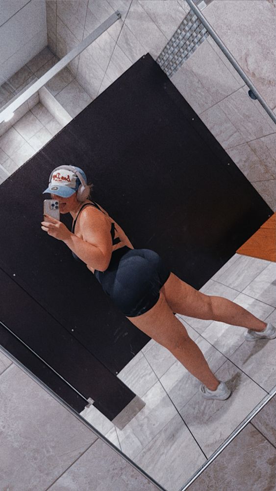 a woman taking a mirror selfie in a locker room stall with her tongue out. she is wearing a black top and bottoms, blue and white sneakers, a blue, white, and red baseball cap, and purple headphones. 