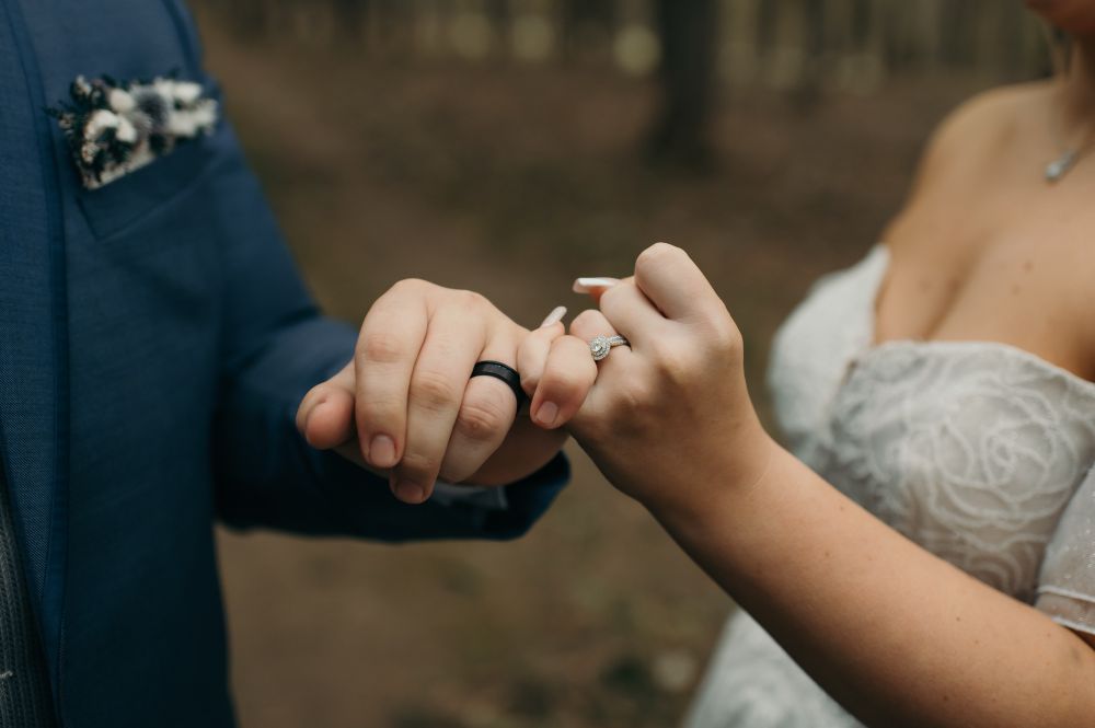 a man in a tux and a woman in a dress standing in the forest, their faces can’t be seen and they have locked pinkies