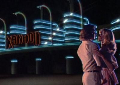Olivia and Michale Beck (the guy from The Warriors) out in front of the rebuilt Xanadu night club/roller rink (?) at night. The building is a classic in the Streamline Moderne style, like something you'd see in a Batman comic, and its swooping embellishments are illuminated with neon blue light.
Fun fact, both me and my wife though this building was a matte painting the first time they showed it, but NO! It was a real building that burned down in the late 80s!