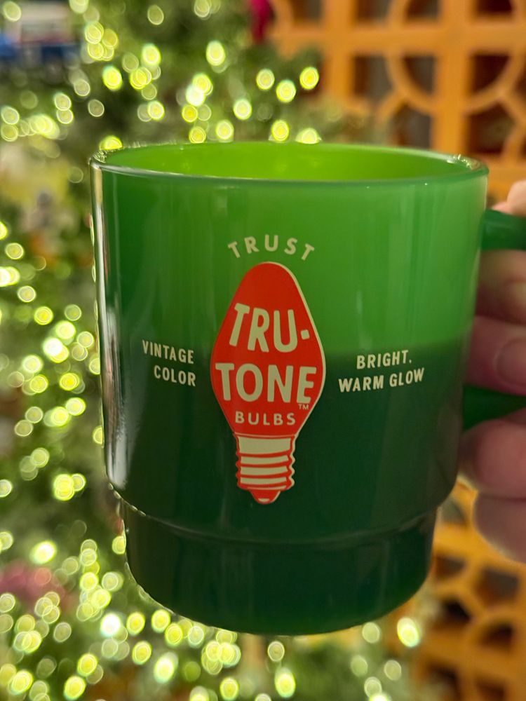 A green glass mug with a red “Tru-tone” C9 Christmas lightbulb on it is in front of a Christmas tree with warm white lights and a breeze block wall. 