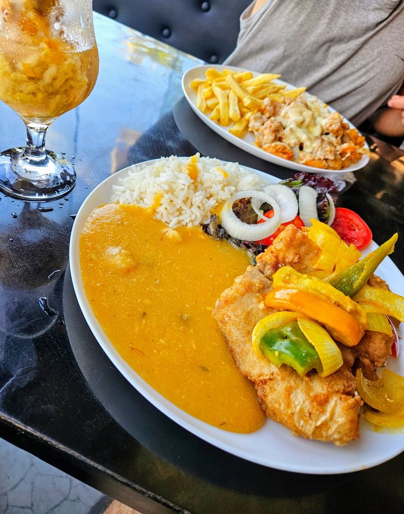 Taça com caipirinha de caju. Prato em primeiro plano com peixe frito, arroz, salada e pirão. Em segundo plano, prato com camarões fritos, arroz e batata frita