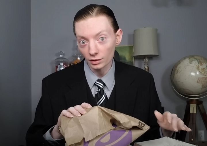 A gaunt, pale man in a suit (definitely a vampire) holding a sack from Taco Bell and looking directly at the camera