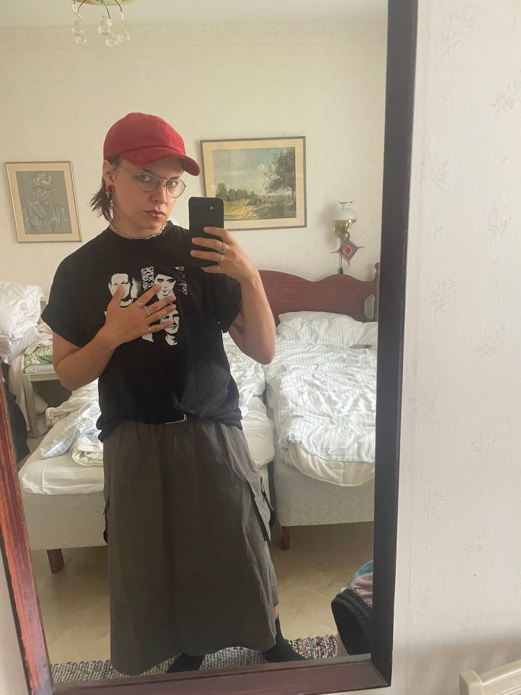 mirror selfie of me wearing a straight long grey-ish skirt with utility pant-vibe pockets + a black border daniel t shirt. i’ve also got a red corduroy cap, red silicone tunnels in my earlobes, and some red eyeshadow underneath my eyes
