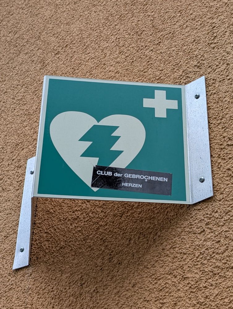 Foto eines Schildes, das einen öffentlichen Defibrillator anzeigt, es ist grün und an einer Hauswand befestigt, darauf ist ein Herz-Piktogramm, durch das ein Blitz geht. Jemand hat einen Aufkleber auf das Schild geklebt mit dem Text "Club der gebrochenen Herzen".