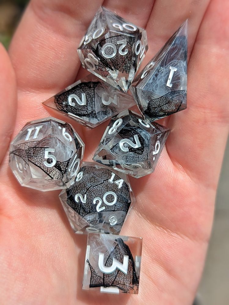 A set of 7 dice. They have white swirls and black skeletal leafs. The numbers are white.