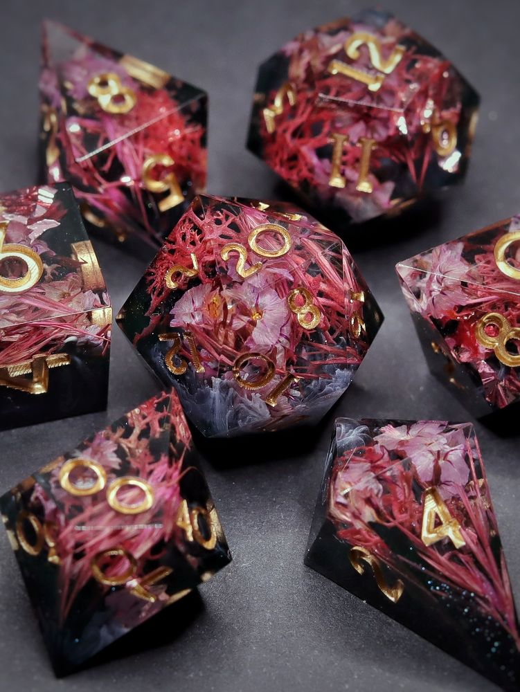 A set of 7 ttrpg dice. They have different pink flowers against a black resin background with swirls. The numbers are gold.