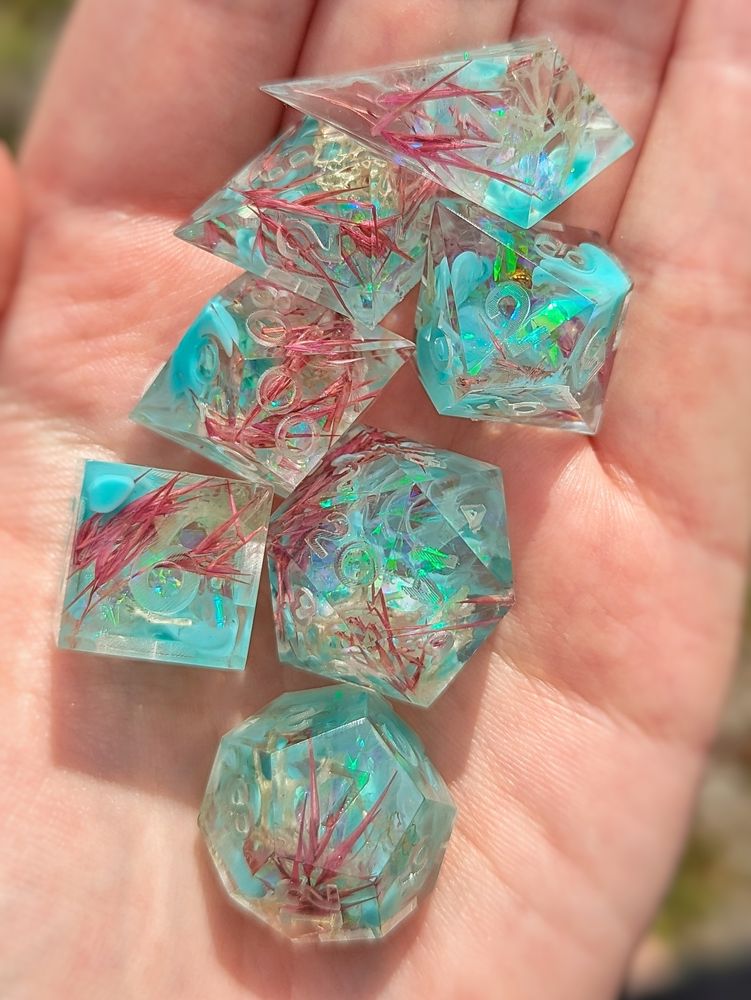 A set of dice. They have teal resin flowing over pink florals and white moss and Holo foil.