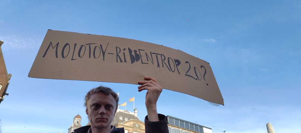 Ein Mensch hält ein Protest-Schild in der Hand, auf diesem steht "Molotov-Ribbentrop 2.0?" zur heutigen Aufteilung der Welt zwischen Trump und Putin. Im Hintergrund ein hellblauer Himmel, mit einigen sehr zarten Wolken 