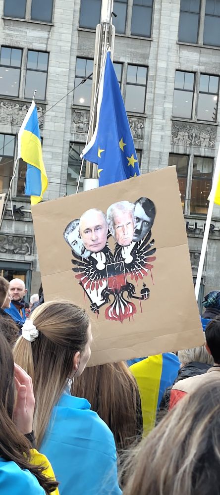Protest-Karton auf einer Demo für die Ukraine, zum 3. Jahrestag der Voll-Invasion Russlands. Auf dem Karton ist in der Mitte ein Wappen-Adler mit zwei Köpfen, rechts Putin, links Trump, als Farbe-Fotos. Hinter jeweils beiden ist, gleichsam lauernd oder als Schatten: rechts hinter Putin, Josef Stalin, hinter Trump Adolf Hitler, beide schwarz-weiss Photos. Alle Köpfe sind sehr sauber ausgeschnitten und zu dieser Kollage zusammen gefügt.