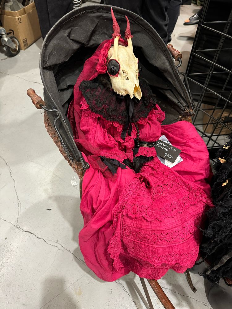 A deer skull in a red and black dress in a stroller with the price tag saying “bebe’s first exorcism”