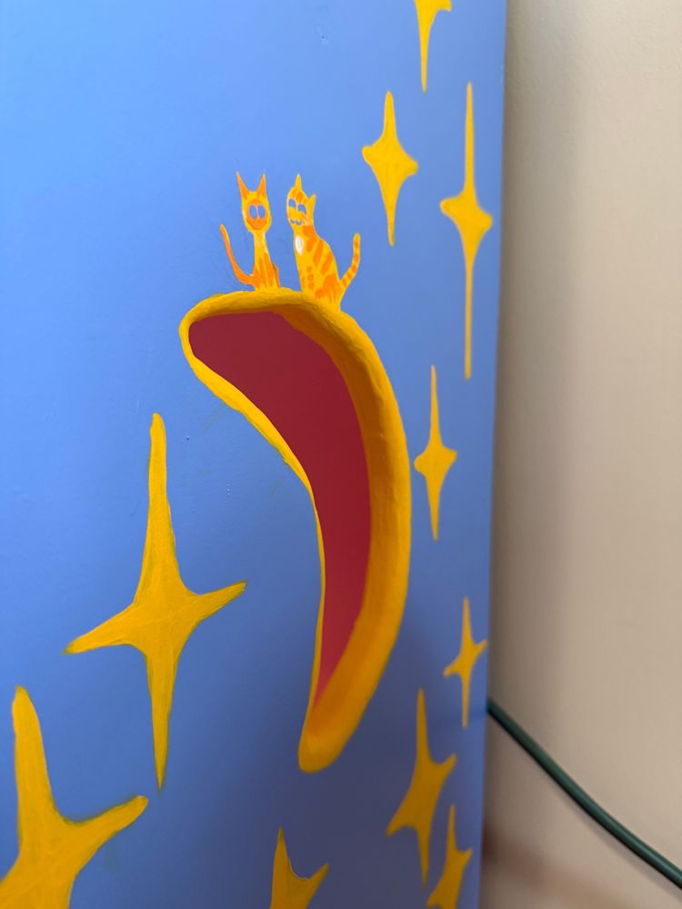 A close up image of one of the moon cut outs on the shout. 2 cats painted in yellow and orange sit on top of it. one is an orange tabby with his eyes closed happily (Tiger), and one small tonkinese cat with big eyes (Lalli) 