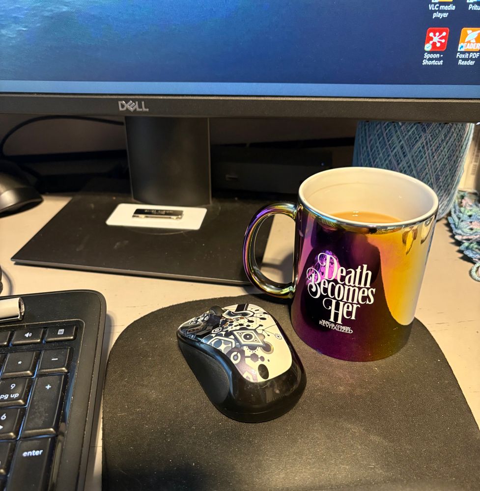 Coffee mug sitting on work desk in front of work computer. Mug is metallic purple with “Death Becomes Her” musical logo.
