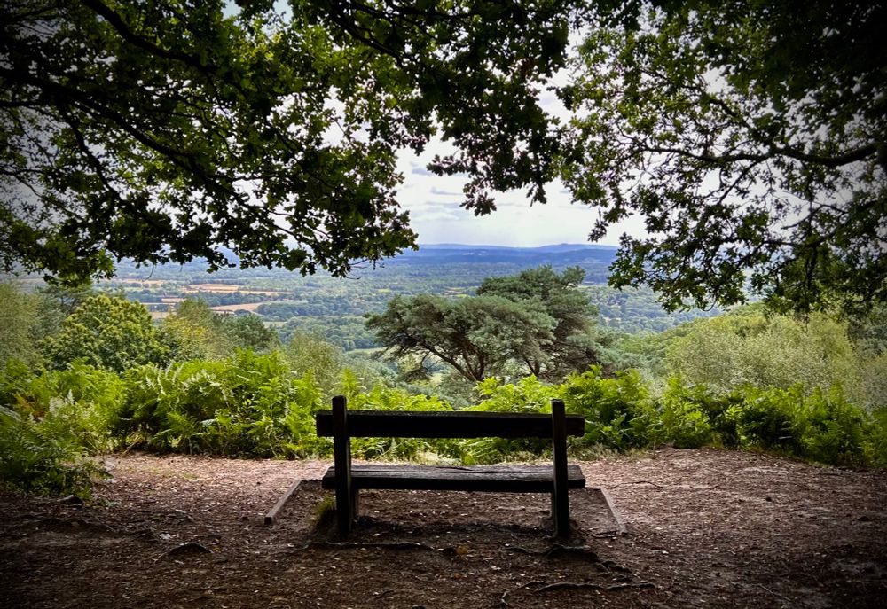 This is a view from near the top of Blackdown. It is the highest point in West Sussex and the South Downs National Park. I believe the hill in the distance is Leith Hill in Surrey, the highest point on the Greensand Ridge.
The photo shows a bench in the foreground with a tree covering and an expansive view of the south western part of Surrey.