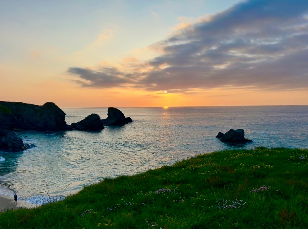 There are dark clouds in the sky. The sun is reflected on the water. A lone figure on the left waits and watches the sunset.