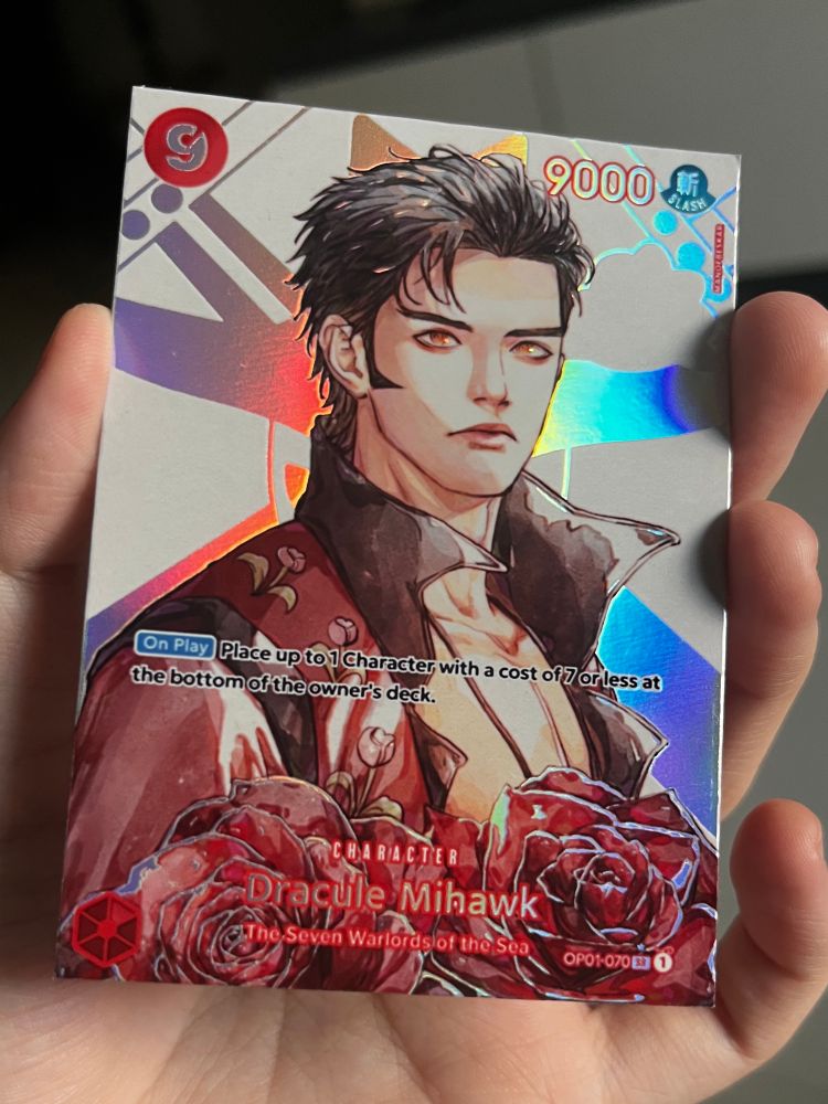 photo of a fanmade custom one piece card. art is of a young mihawk with two red and black roses covering his chest and going off-card. there is a holographic effect of his official jolly roger. the art has a watercolour effect.