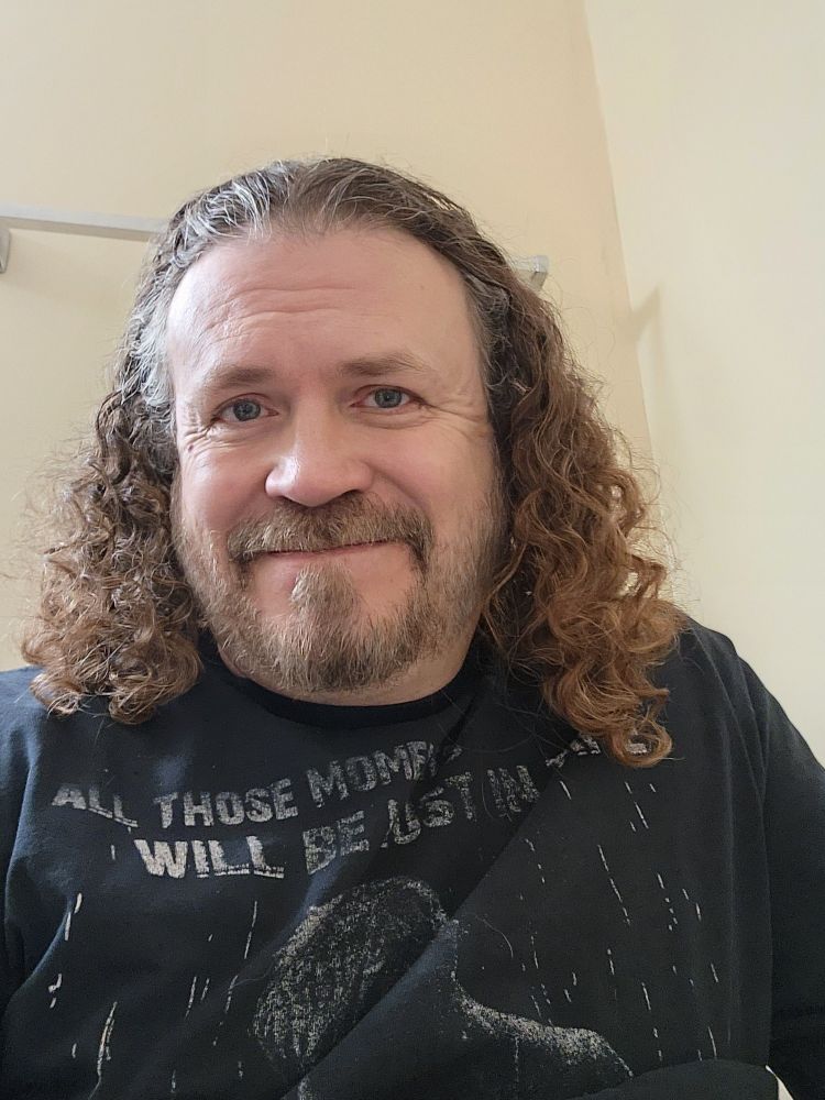 A shrugging Caucasian man with long curly brown hair and a short brown beard, wearing a black shirt that reads "All those moments will be lost in time like tears in rain", a quote from the film Blade Runner. 
