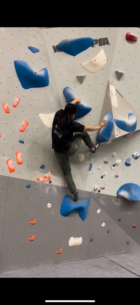 A woman with long black hair climbing on the wall in a climbing gym. The route has huge and slippery blue holds that are mostly slopers. She is leaning back and locking her shoulders in order to keep the position and swift the weight on the other side. She is hanging on the holds and lifting her right leg on the volume close to her right hand.