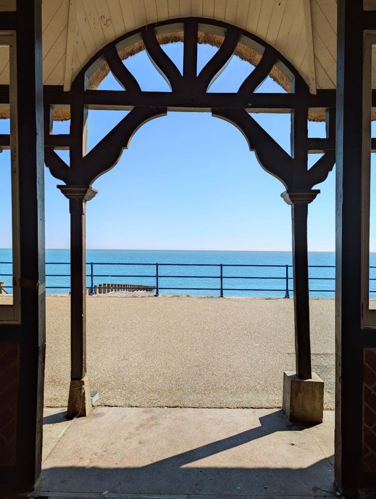View from inside a dark wooden shelter looking out through an ornate archway onto a sunny seaside promenade, with the calm blue sea and a clear sky in the background.