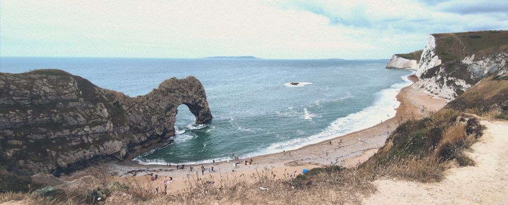 A panoramic shot captures the iconic Durdle Door, a natural limestone arch, on a shingle beach in Dorset, England. The arch juts out into the calm, blue-green sea, with gentle waves lapping the shore. White cliffs frame the right side of the image, stretching into the distance under a partly cloudy sky. Scattered beachgoers are visible on the shingle beach, and a distant headland is faintly visible on the horizon.
