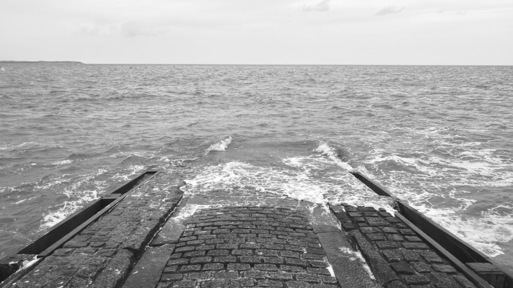 A black and white image of a brick causeway jutting into a frothy sea. 