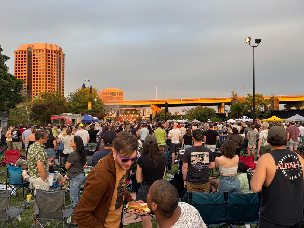A crowd of concertgoers with an outdoor stage in the distance, near sunset. 
