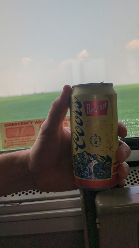 A hand holds a can of Coors Banquet aboard a train looking out the window into a green soybean field.