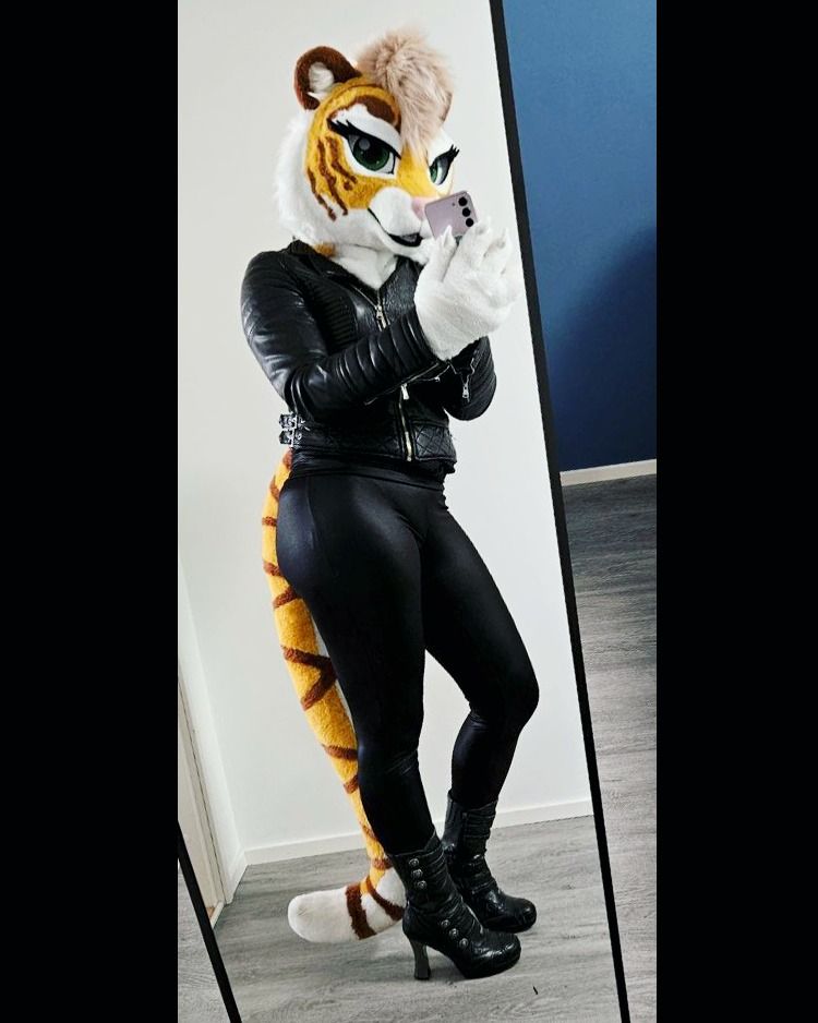 A female tiger partial fursuit posing in front of a mirror taking a selfie with a lilac phone. She's wearing a black leather jacket, black leather leggins and black high heeled leather boots.