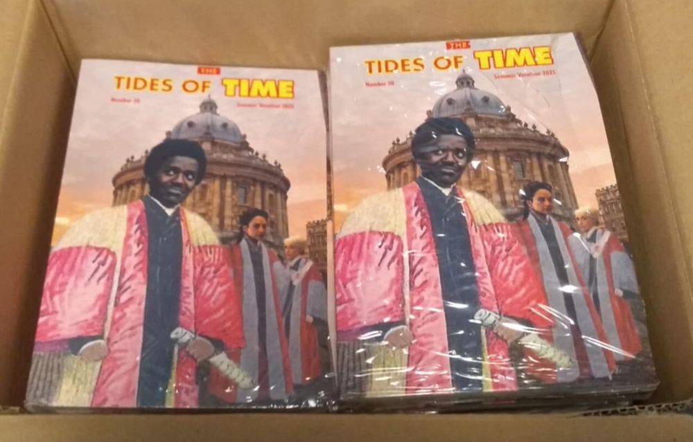 Two piles of a printed magazine in a box. The cover shows the Fifteenth Doctor Who in the gown of an Oxford DMus, with the two incarnations of the Rani played by Archie Panjabi and Anita Dobson in the gowns of Oxford DScs, in front of the Radcliffe Camera.