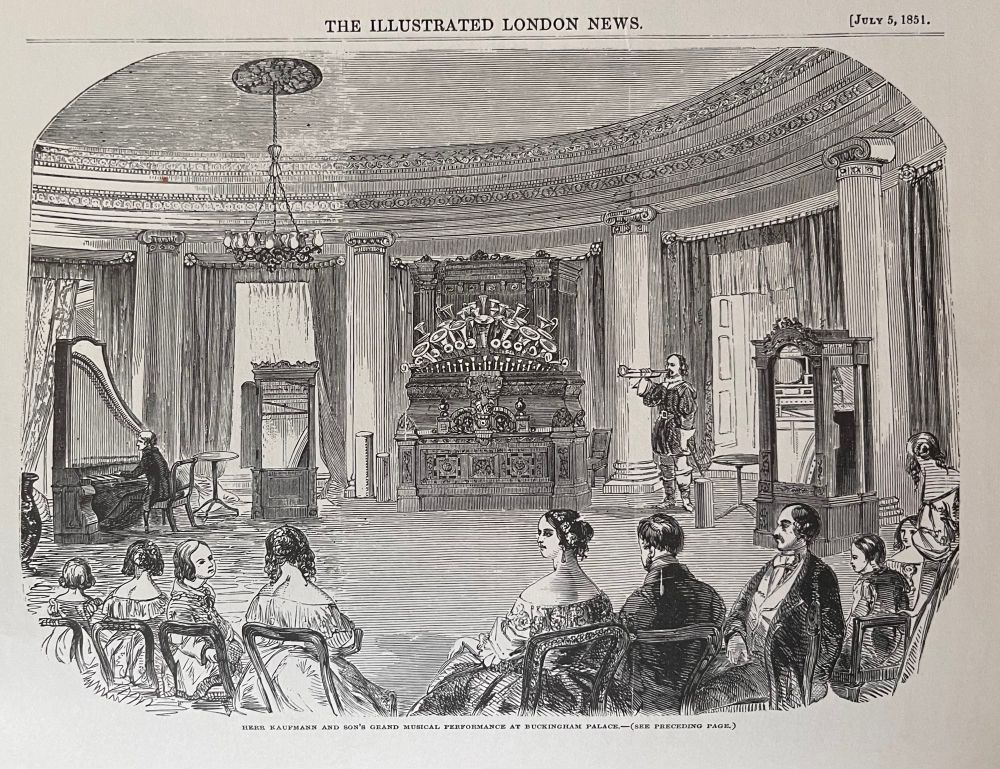 Victorian-era engraving from The Illustrated London News, dated July 5, 1851, showing an elaborate musical performance at Buckingham Palace. At the center is a large orchestrion—an ornate mechanical instrument with multiple brass horns and decorative carvings. A formally dressed man operates the orchestrion while another stands nearby playing a wind instrument, possibly a trumpet or cornet. A small audience, dressed in 19th-century attire, sits in rows of chairs watching the performance. The grand room features tall columns, chandeliers, draped curtains, and high ceilings, emphasizing its regal setting. The caption reads: “Herr Kaufmann and Son’s Grand Musical Performance at Buckingham Palace.”