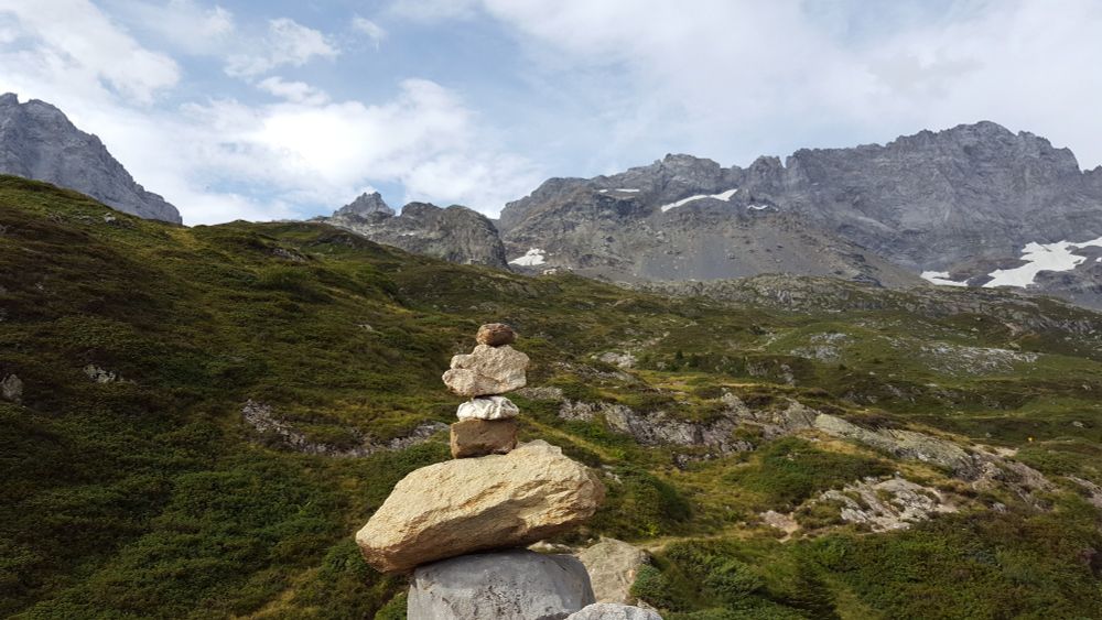 The background is steep, grey, jagged mountain tops giving way to still steep green mountains with a barely visible foot path and little cabin. In the foreground are a bunch of differently shaped stones put on top of each other.
