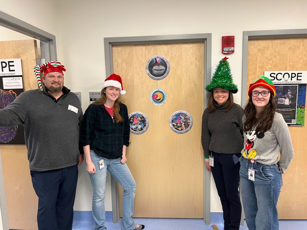 From left to right, Vizgen engineer Nick wearing and elf hat, Dr. Christina Baer in a christmas tree hat, Jill McConnell program manager in a santa hat, Molly Martin post doc in an elf hat all in front of the Merscope room door decorated with the CSP logo and portholes with sea creatures in party hats. 